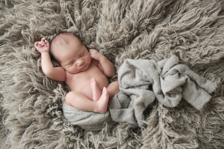 newborn baby laying on a gray blanket with hands over his head