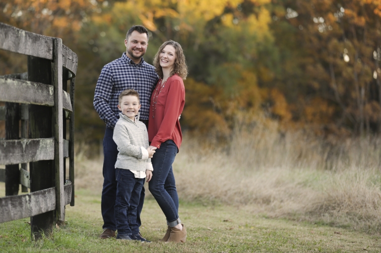 Family of three standing up against a fence with beautiful fall colors behind them